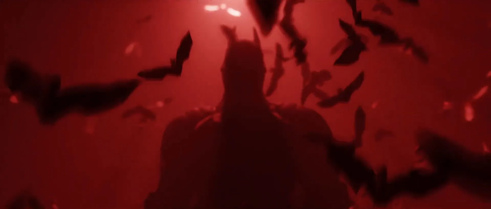 Batman's dark silhouette standing in deep red light beneath a spotlight as a swarm of bats scatters around him.